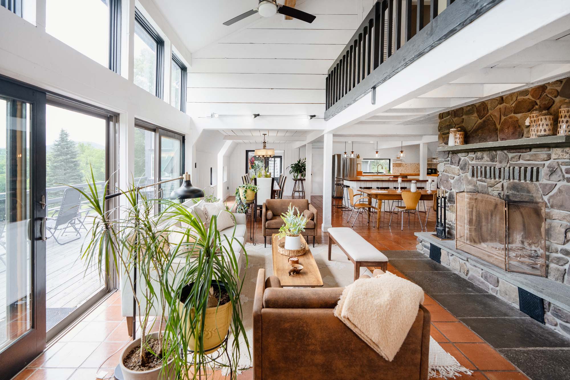 Spacious, well-lit living room and dining area with large windows and glass sliding doors on the left, leading to an outdoor deck with chairs. Inside, there are brown armchairs, a white sofa, a wooden coffee table with plants, and a white bench. A stone fireplace is on the right, and a wooden dining table with chairs is in the background. Ceiling fans and potted plants are also visible.