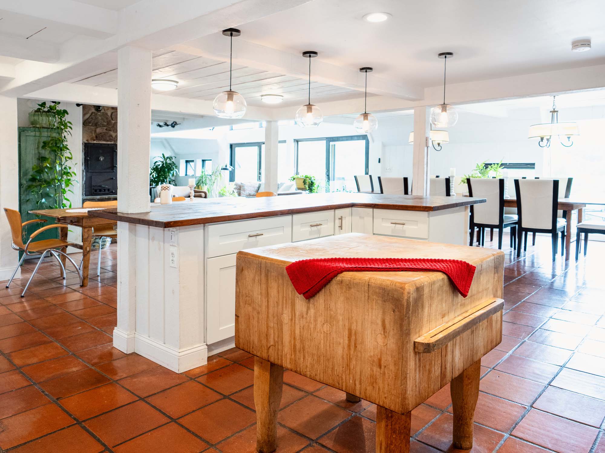 Spacious, bright kitchen and dining area with terracotta tile flooring. A large wooden butcher block with a red cloth on top is in the foreground. Behind it is a white kitchen island with wooden countertops and drawers. Hanging pendant lights are above the island. In the background, there are dining tables with chairs, potted plants, and large windows letting in natural light.