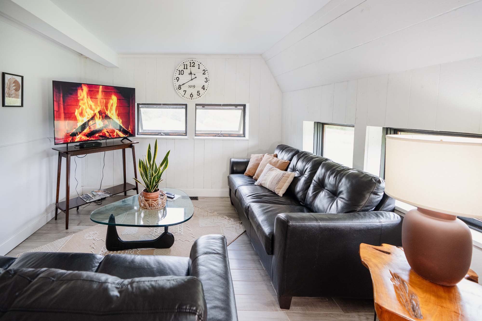 Modern living room with two black leather sofas, a glass coffee table with a potted plant, and a wooden side table with a lamp. A flat-screen TV on a wooden stand displays a fireplace scene. The walls are white with two small windows below a round clock showing 11:40. The floor has light-colored wood or tile flooring with a beige rug.