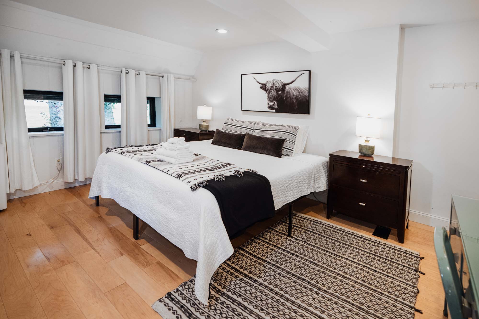 Bedroom with a large bed covered in white bedding and a black and white patterned throw. Two dark brown rectangular pillows and two white pillows with stripes rest against the headboard. On the bed are several folded white towels. Two dark wood nightstands flank the bed, each with a lamp. A black and white animal picture hangs above the bed. The floor is light wood with a black and white patterned rug partially under the bed. There are three small windows with white curtains on the left wall.
