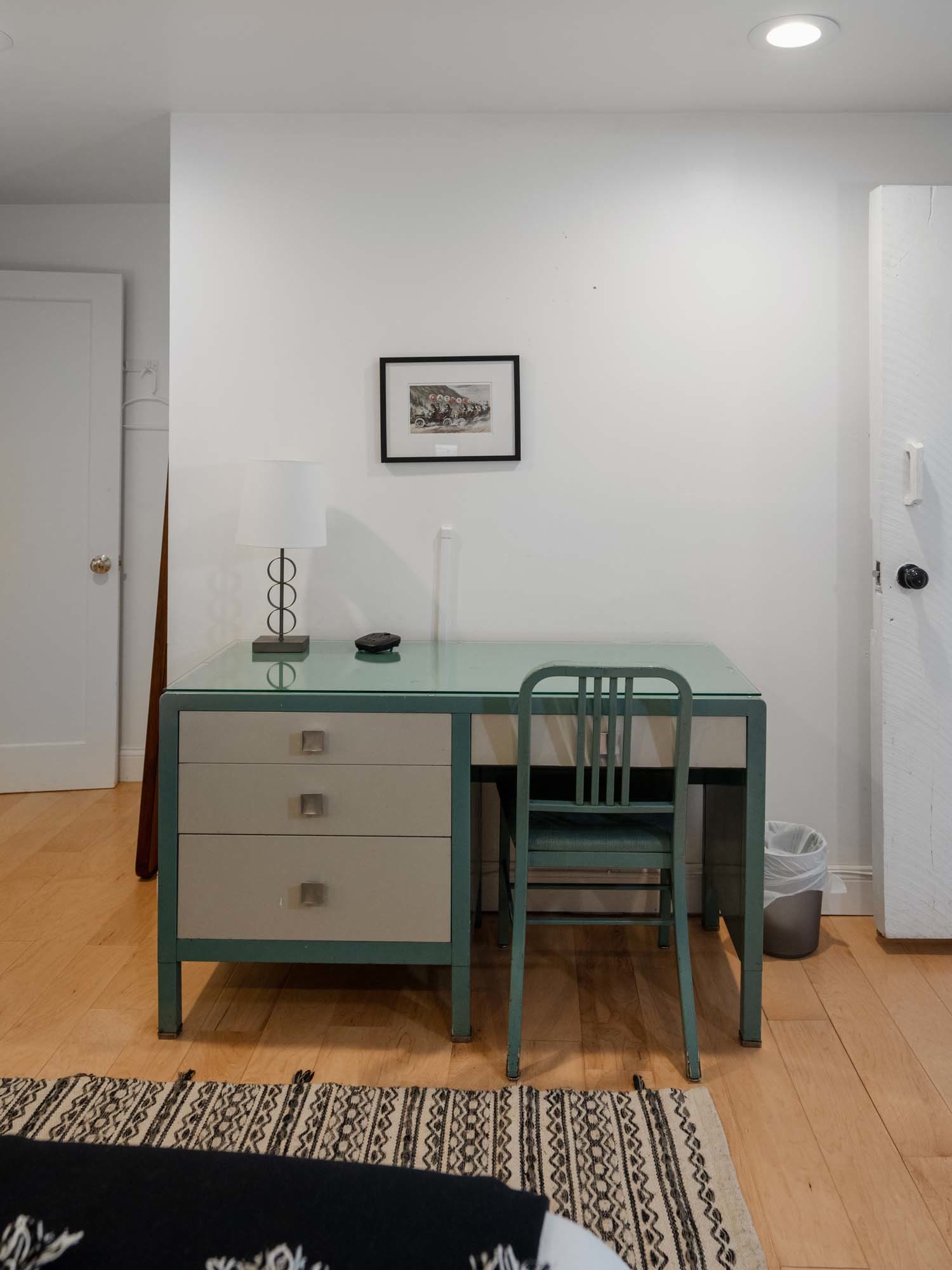 Minimalist room with a vintage-style desk and chair set against a plain white wall. The desk has three drawers on the left side and a glass top. On the desk, there is a table lamp with a white lampshade and a small black object. Above the desk hangs a small framed picture. The floor is wooden with a patterned rug partially visible. Two doors are visible on either side of the desk, and a small trash can is placed near the right door.