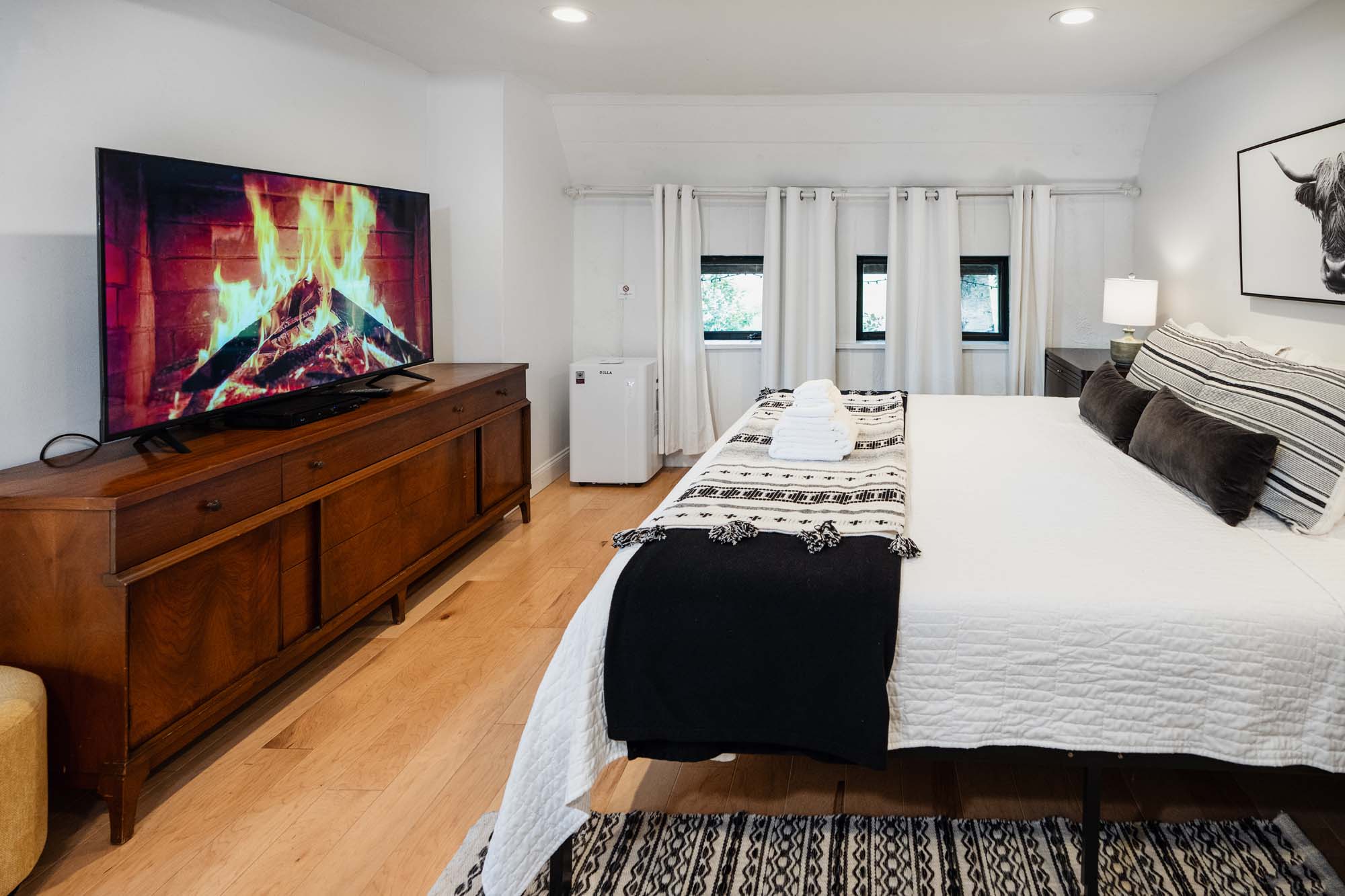 Bedroom with a large bed covered in white bedding and black and white patterned blankets. Three dark pillows are on the bed. A stack of white towels is placed on the blanket at the foot of the bed. To the left, a wooden dresser holds a TV displaying a fireplace with burning logs. The room has light wooden flooring, a patterned rug, two small windows with white curtains, a white mini fridge, and a nightstand with a lamp and a framed picture of an animal on the wall.
