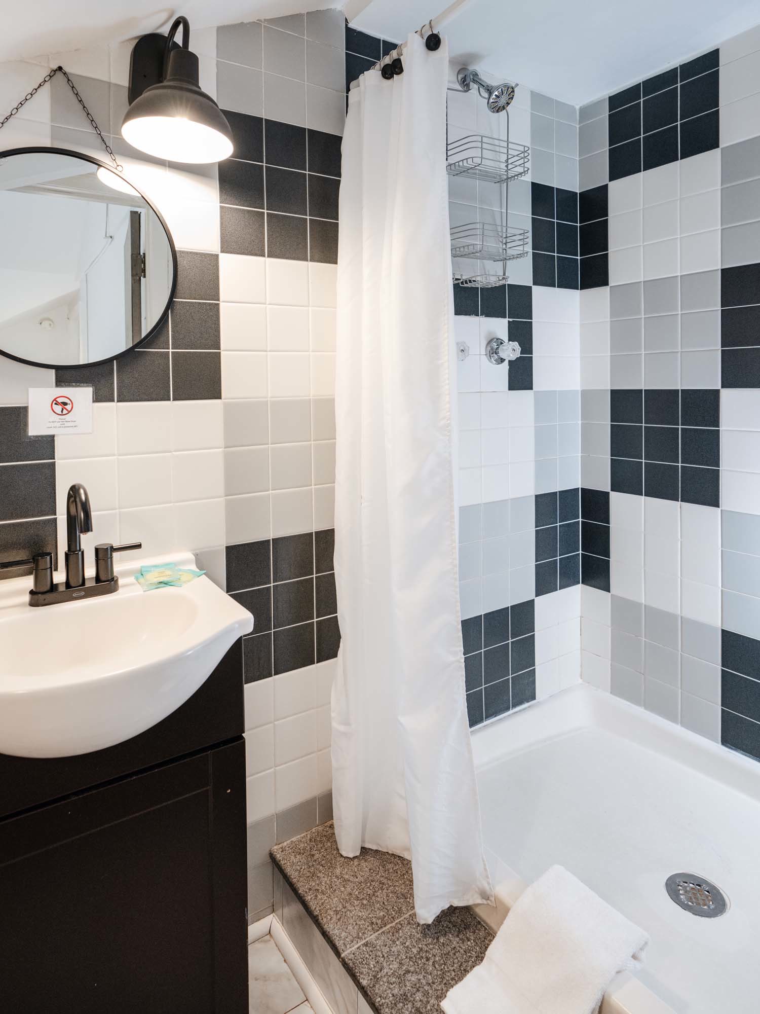 Bathroom corner with a shower area and a sink. The shower has a white curtain, a silver showerhead, and metal racks attached to the tiled wall. The tiles are arranged in a pattern of black, white, and gray squares. The sink is white with a black faucet and is mounted on a black cabinet. Above the sink, there is a round mirror with a black frame and a black wall-mounted light fixture. A white towel is draped over the edge of the shower base.