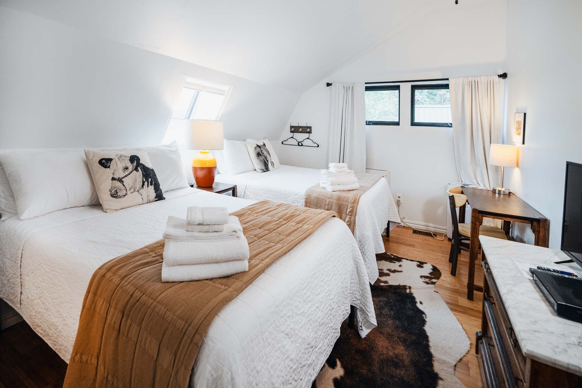 Bright bedroom with two beds, each covered with white bedding and brown throws. Both beds have pillows with cow illustrations. On each bed, there are neatly folded white towels. Between the beds is a nightstand with a lamp featuring a yellow and orange base. The room has light wooden floors, a cowhide rug, a window with white curtains, a desk with a chair and lamp, and a TV on a marble-topped dresser.