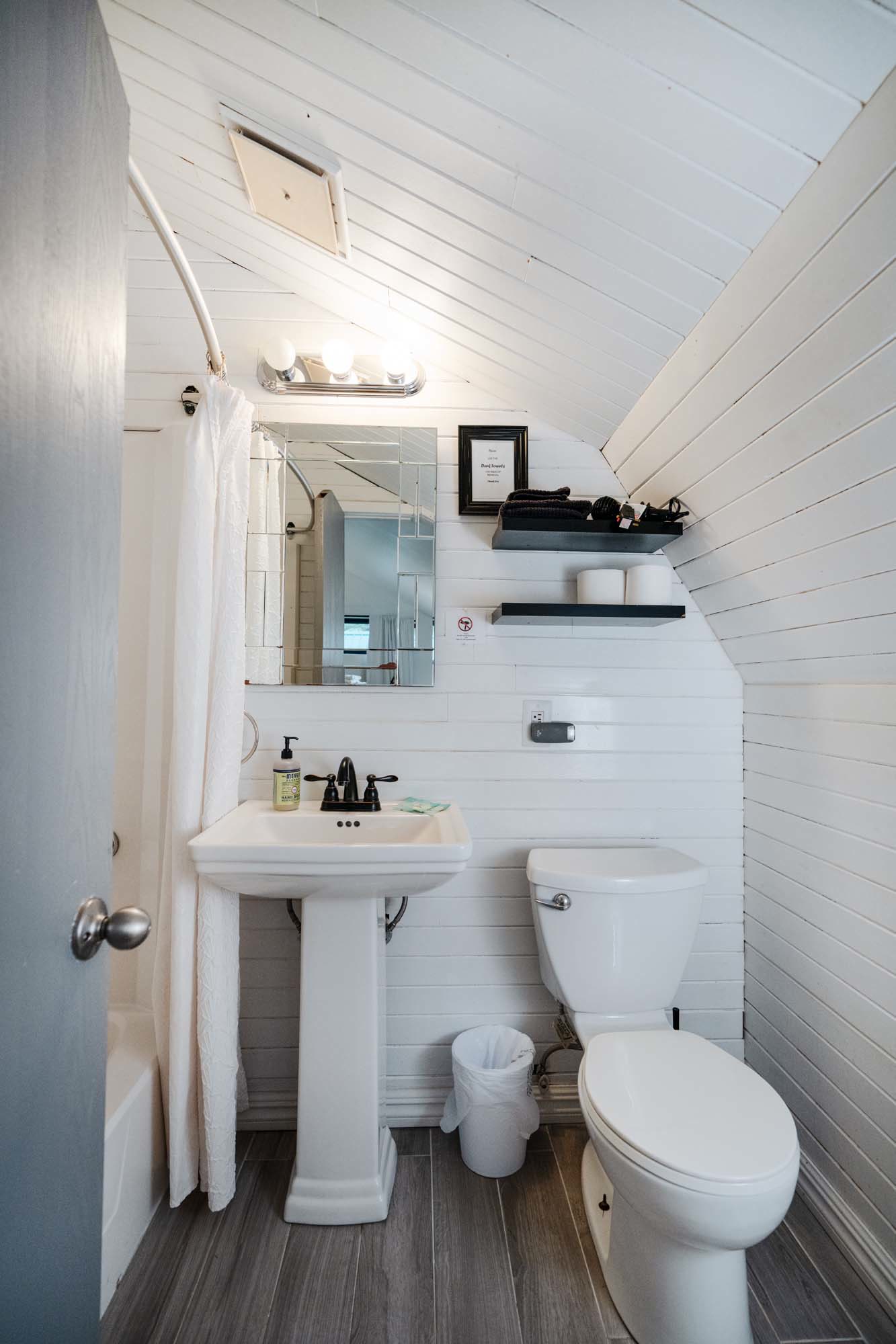 Small bathroom with white wooden panel walls and a slanted ceiling. It features a white pedestal sink with a black faucet, a wall-mounted mirror above the sink, and a white toilet next to it. Two black shelves are mounted on the wall above the toilet, holding black towels, toiletries, and two rolls of toilet paper. A small trash bin is placed between the sink and toilet. A shower curtain is partially visible on the left side, and the floor has gray wooden planks.