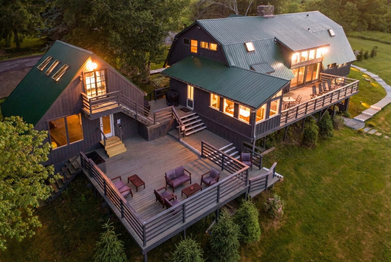 Large, two-part house with dark exterior walls and green metal roofs. The house has multiple windows, some illuminated from inside. There are two connected sections with stairs and railings leading to a spacious wooden deck furnished with several purple cushioned chairs and small tables. The house is surrounded by green grass and trees, with a paved walkway visible on the right side.