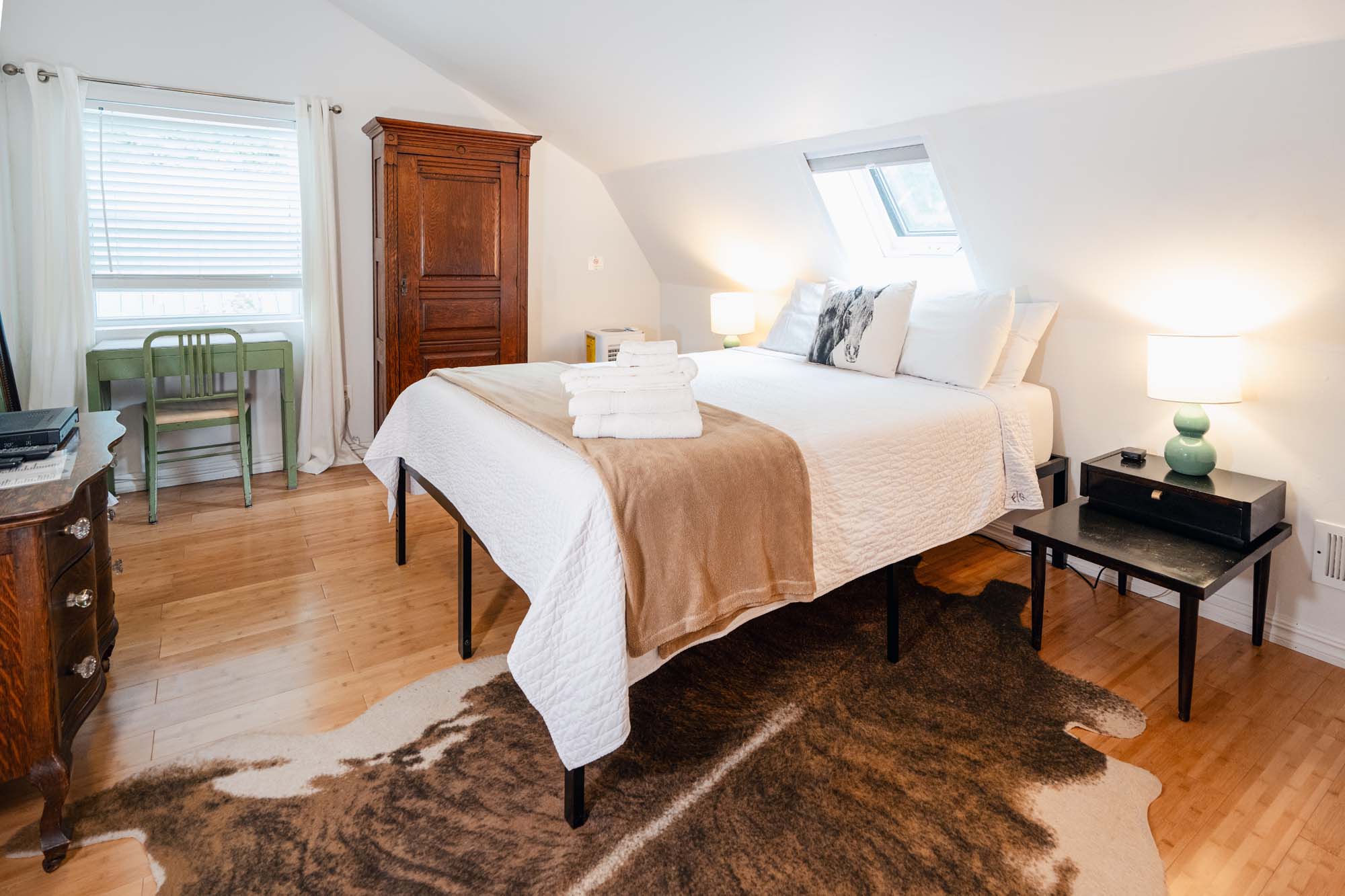 Bedroom with a large bed covered in white bedding and a beige throw blanket. There are stacked white towels on the bed and pillows, including one with a horse image. A brown and white rug is on the wooden floor beneath the bed. The room has a wooden wardrobe, a green desk with a chair, two bedside tables with lamps, and a window with blinds and curtains.