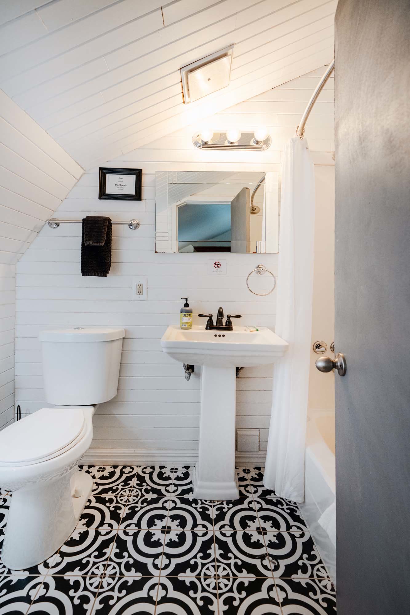 Small bathroom with white wooden paneled walls and a sloped ceiling. There is a white toilet on the left and a white pedestal sink in the center with a black faucet. Above the sink is a rectangular mirror with a light fixture containing three bulbs. A black towel hangs on a rod to the left of the mirror. The floor has black and white patterned tiles. A white shower curtain is partially visible on the right side of the image.