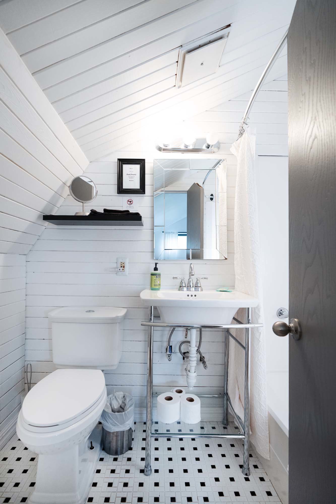 Small bathroom with white wooden paneled walls and a slanted ceiling. It features a white toilet with a silver trash bin beside it. A white sink with a chrome frame holds three rolls of toilet paper underneath. Above the sink is a rectangular mirror with a light fixture above it. A black shelf on the wall holds a round mirror and folded towels. A shower curtain is partially visible on the right side.