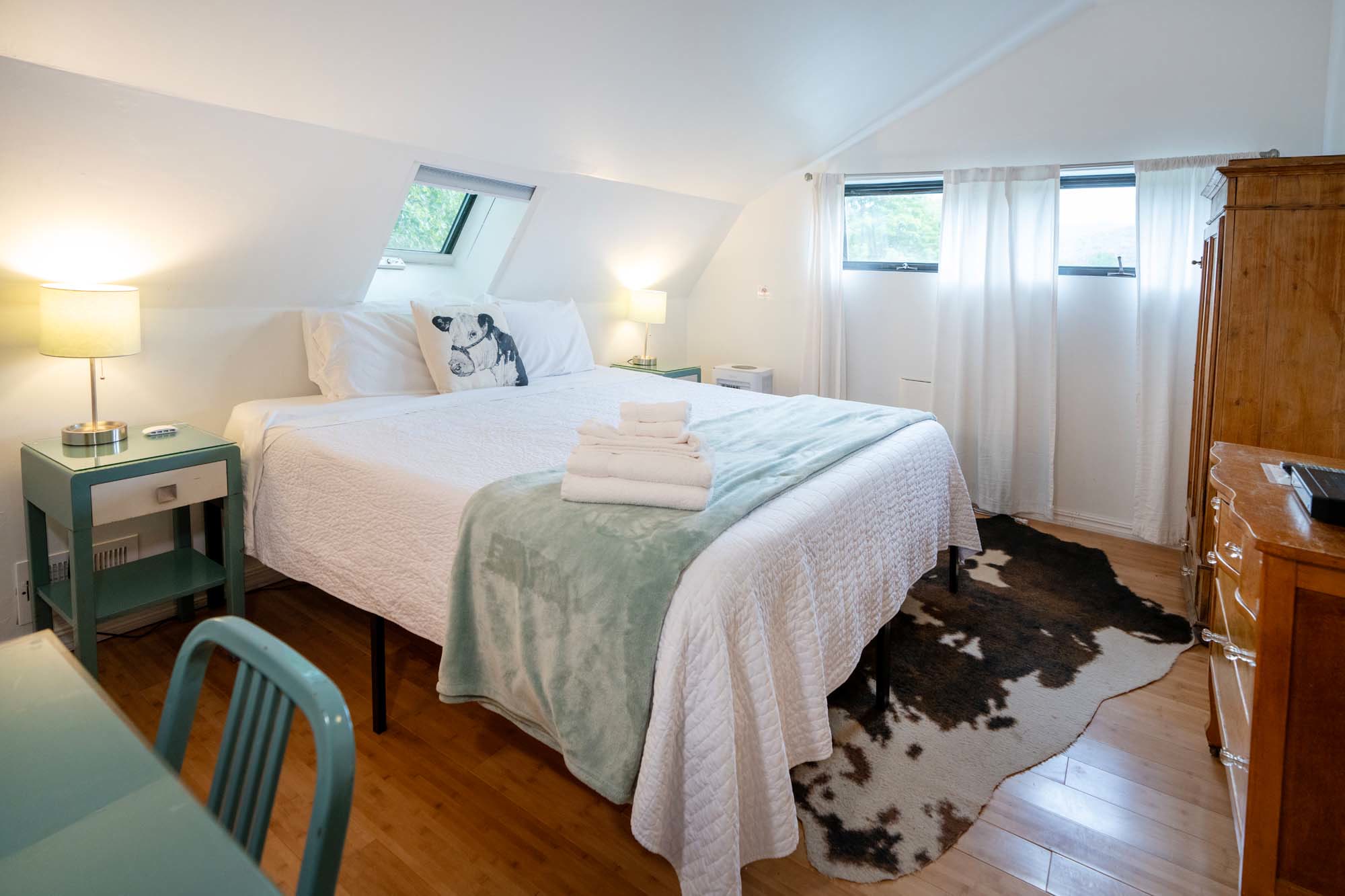 Bedroom with a large bed covered in a white quilt and a light blue blanket at the foot. On the blanket, there are folded white towels. The bed has white pillows, one featuring a black and white dog illustration. Two bedside tables with lamps flank the bed. The room has wooden floors with a brown and white cowhide rug. A wooden dresser is on the right, and two windows with white curtains are on the far wall.