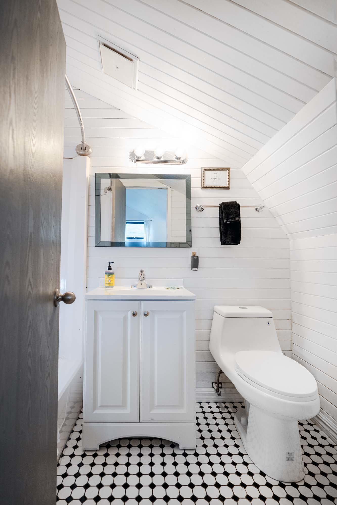 Small bathroom with white wooden panel walls and a slanted ceiling. There is a white vanity with a sink and a bottle of yellow liquid soap on the left. Above the vanity is a rectangular mirror with a black frame and a three-bulb light fixture. To the right of the vanity is a white toilet. A black towel hangs on a towel bar above the toilet. The floor has black and white hexagonal tile patterns. A partially open wooden door is on the left side.