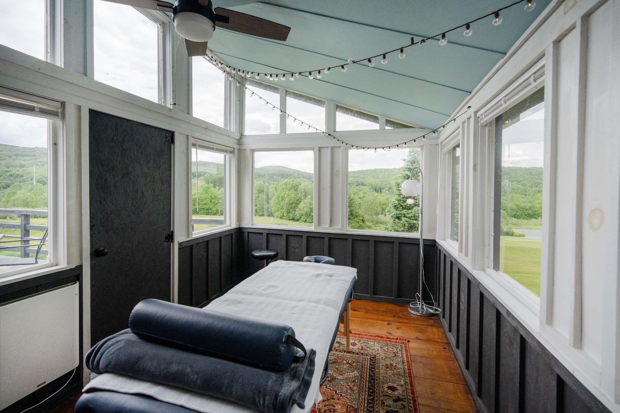 Small enclosed room with large windows on three sides, offering a view of green trees and hills outside. Inside, there is a massage table with a white sheet and a dark blue bolster pillow on top. The floor is wooden with a patterned rug underneath the table. A black door is on the left wall, and a string of small lights is hung across the ceiling. A floor lamp with a round white shade stands near the windows on the right side.