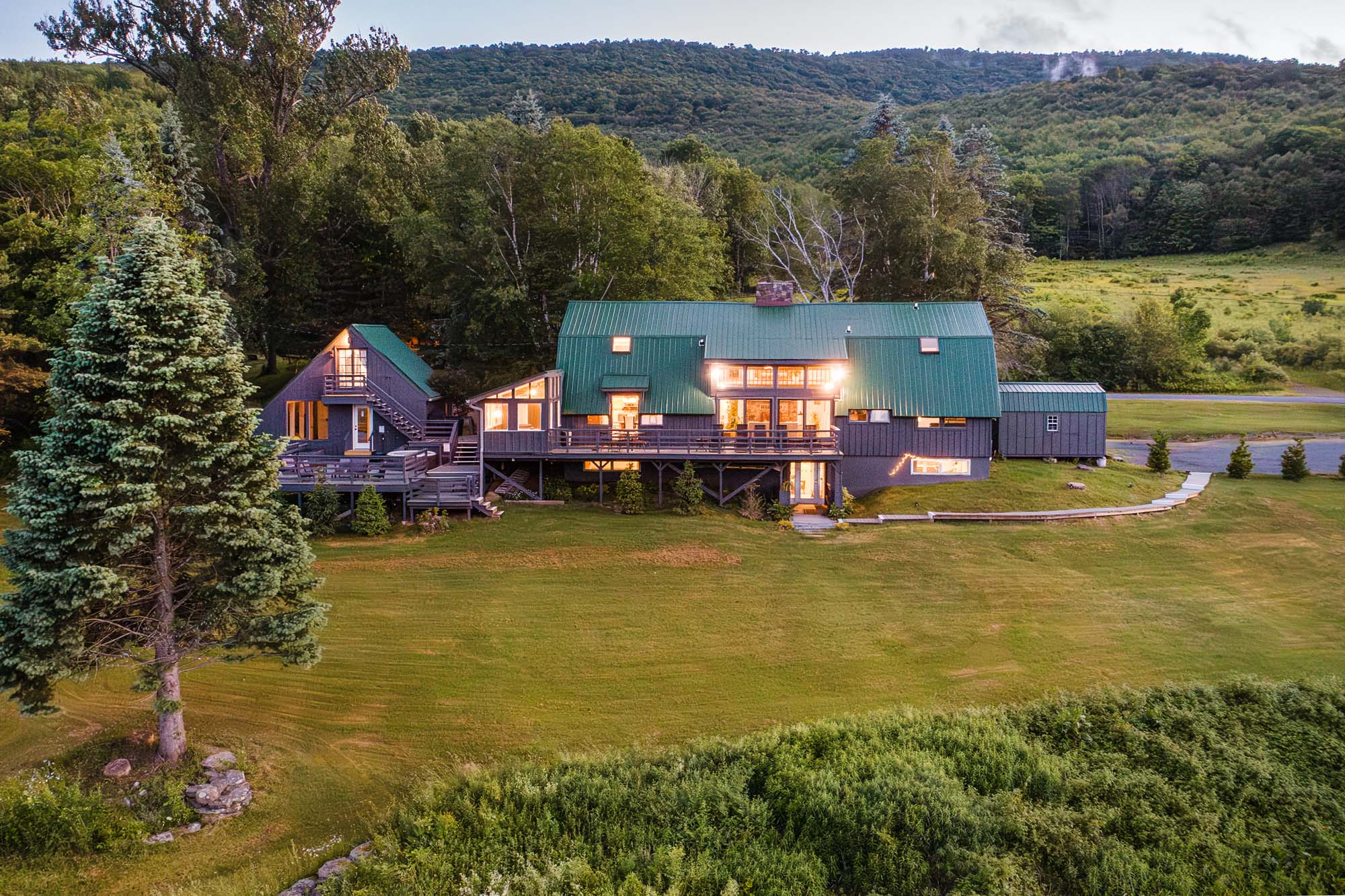 Large house with a green roof and multiple sections, including a main building and a smaller adjacent structure. The house has multiple windows, some lit from inside, and a wooden deck with railings. It is situated on a well-maintained grassy lawn with a tall evergreen tree in the foreground. The background features dense trees and hills under a cloudy sky.