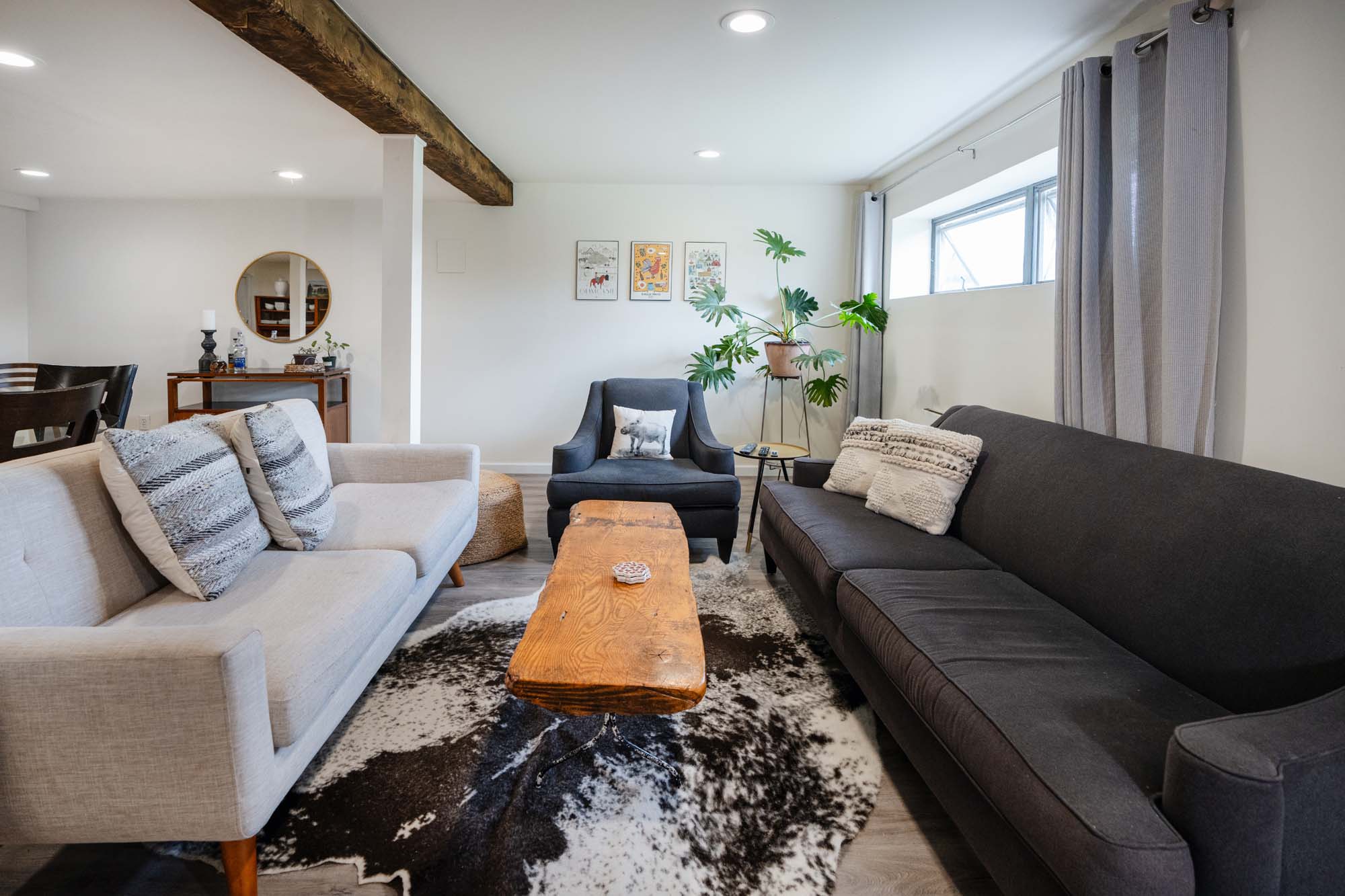 Living room with a light gray sofa on the left, a dark gray sofa on the right, and a dark gray armchair in the center. A wooden coffee table sits on a black-and-white patterned rug. Behind the armchair is a large green plant in a pot on a metal stand. Three framed pictures hang on the white wall. A window with gray curtains is on the right wall.