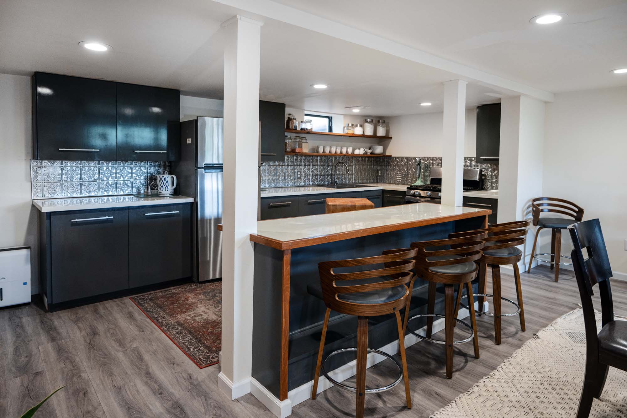 Modern kitchen with dark cabinets and a light-colored countertop. There are three wooden bar stools with metal footrests lined up at the kitchen island, which has a white countertop with a wooden edge. The kitchen has stainless steel appliances, including a refrigerator and a stove. The backsplash features a patterned metallic tile. Open shelves hold jars and cups. The floor is wood-like, and there is a patterned rug near the refrigerator. The ceiling has recessed lighting.
