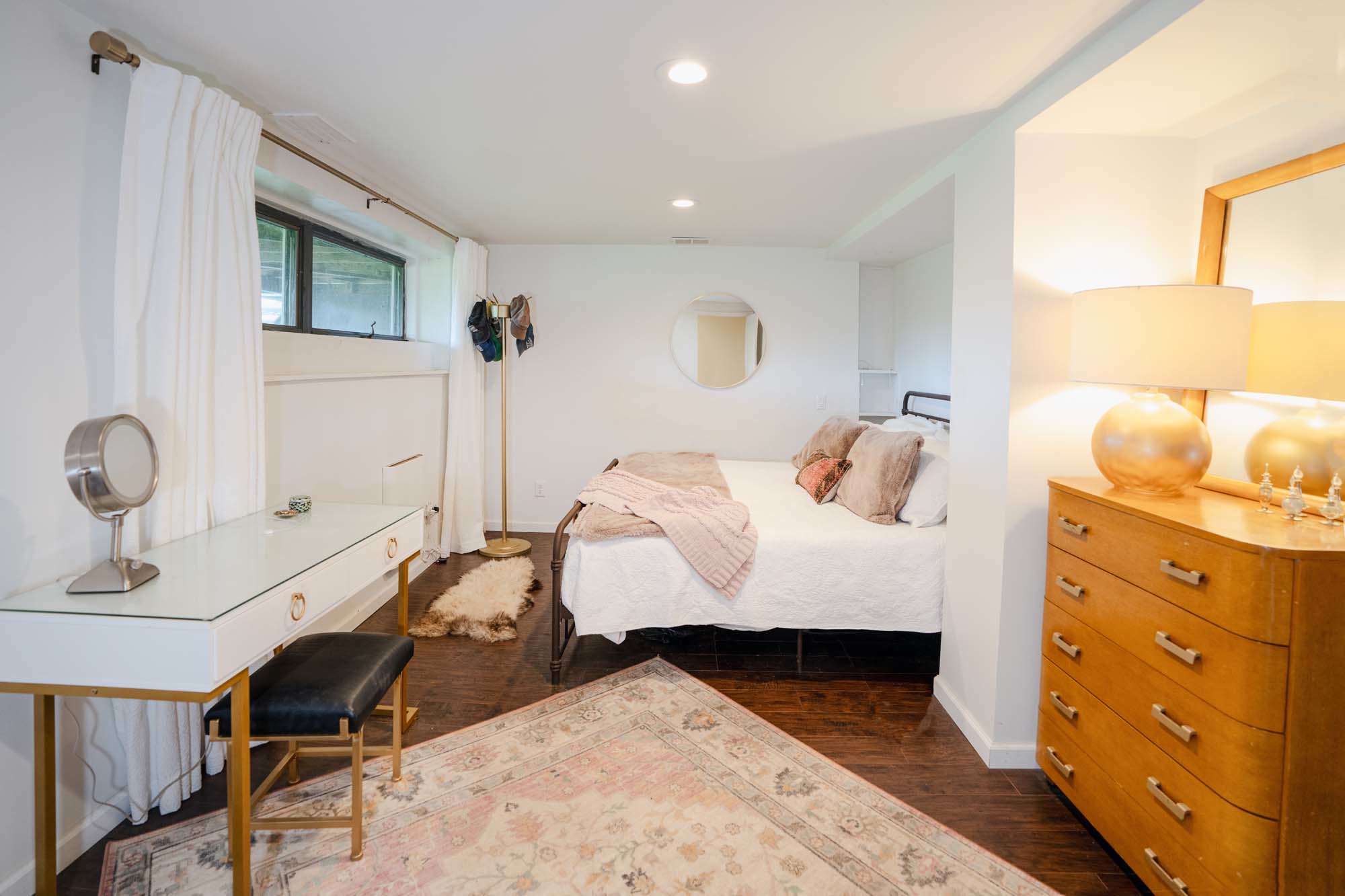Bedroom with white and pink color scheme. It has a bed with white bedding, pink and beige pillows, and a pink throw blanket. To the right is a wooden dresser with a large mirror and a round lamp on top. On the left is a white desk with a small round mirror and a black cushioned stool. A fluffy beige rug is on the dark wooden floor, along with a larger patterned rug. A coat rack with hanging items is near a rectangular window with white curtains.