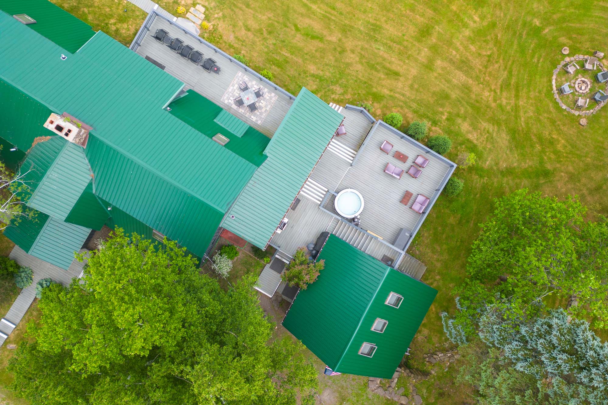 Aerial view of a green-roofed building with multiple sections and outdoor decks. One deck has several lounge chairs and a table with an umbrella, while another deck has seating furniture arranged around a circular hot tub. Surrounding the building is a grassy area with trees and a circular fire pit with chairs placed around it.