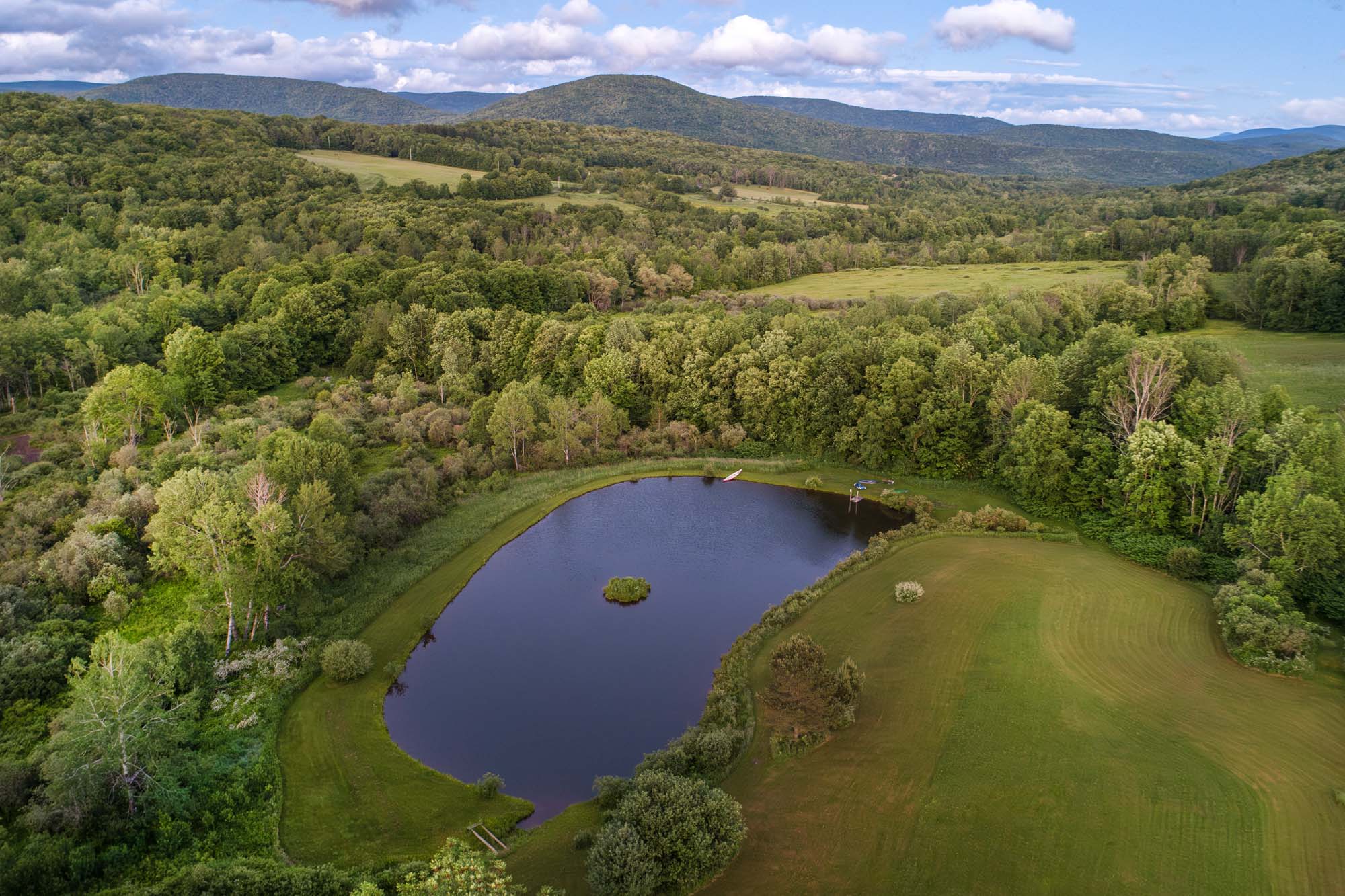 Aerial view of a small, oval-shaped pond surrounded by green grassy areas and dense forest. A small island is visible in the center of the pond. There is a dock with a small boat and a canoe near the pond’s edge. Rolling hills and a mountain range extend into the background under a partly cloudy sky.
