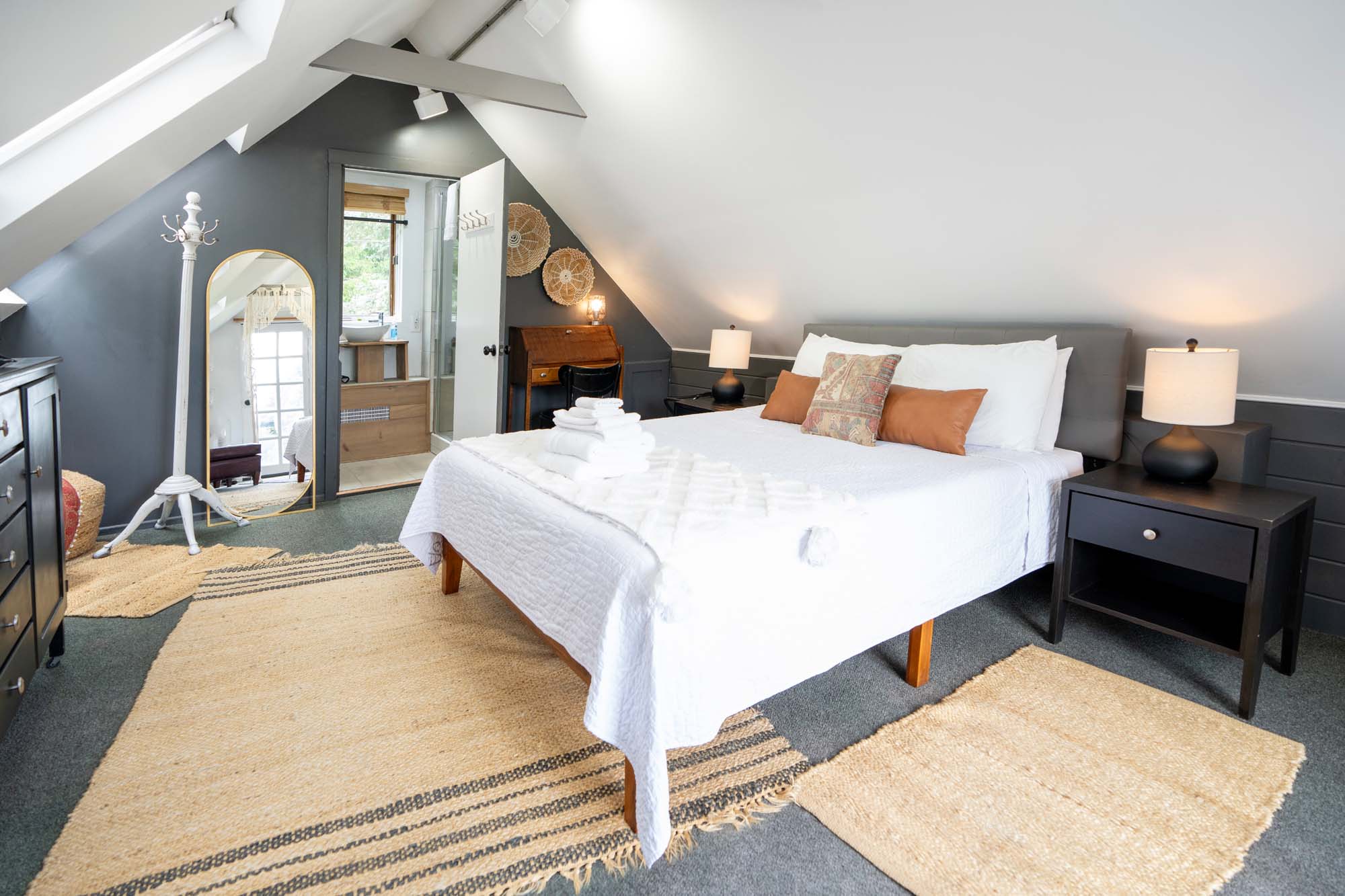 Cozy attic bedroom with slanted ceilings. A bed with white bedding and three pillows (two brown, one patterned) is centered. Two black nightstands with lamps flank the bed. A wooden dresser is on the left. There are beige rugs on the gray carpeted floor. A full-length mirror and a white coat rack stand near an open door leading to a bathroom.