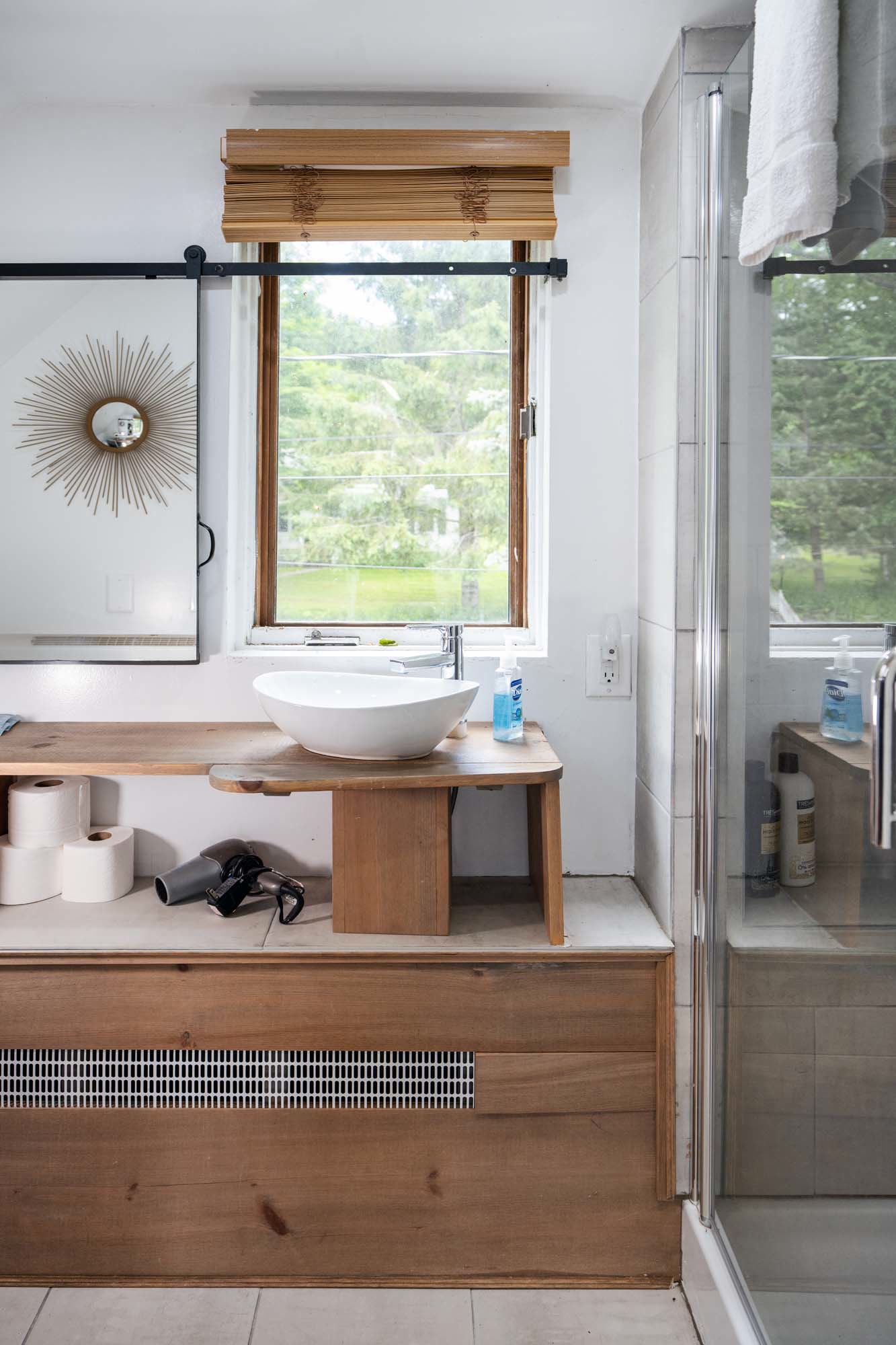 Bathroom vanity area featuring a wooden countertop with a white vessel sink and a chrome faucet. To the right of the sink is a bottle of hand soap. Below the counter are several rolls of toilet paper and a hairdryer. Behind the sink is a window with a bamboo blind partially rolled up. To the right is a glass shower door with shampoo bottles visible inside. A towel hangs on a bar above the shower door.