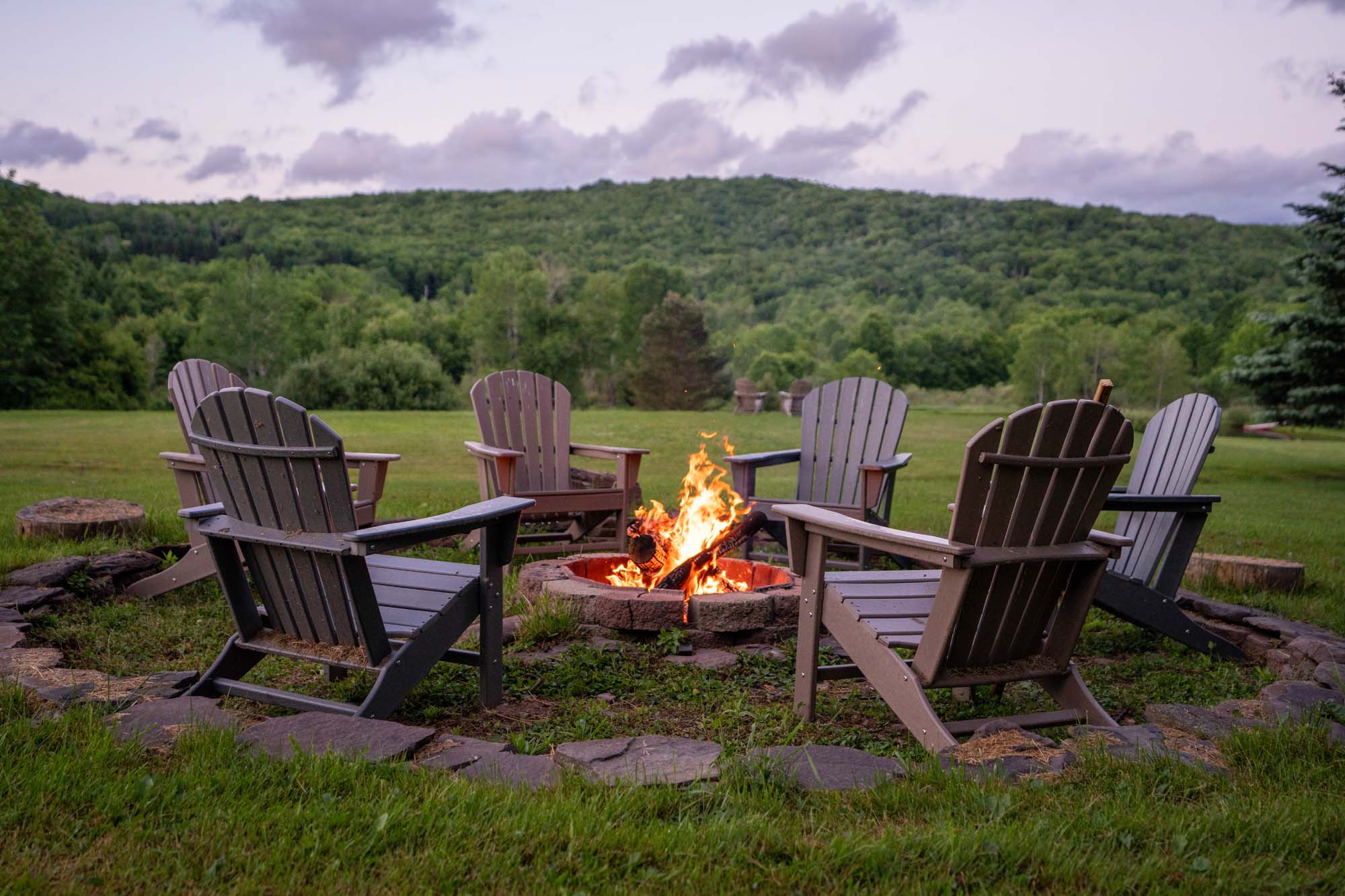 Circular outdoor fire pit with a fire burning inside it, surrounded by six Adirondack chairs arranged evenly around the fire. The setting is a grassy area with a backdrop of dense green trees and a forested hill under a partly cloudy sky during dusk or early evening.