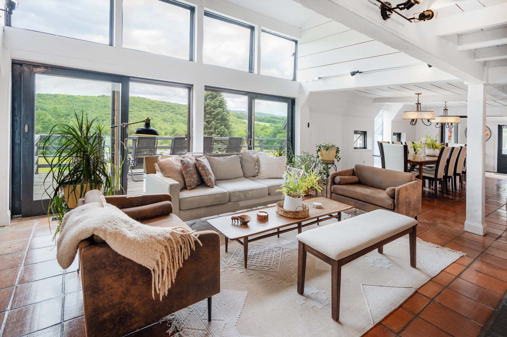 Spacious living room with large windows and sliding glass doors offering a view of green hills outside. The room has a beige sofa, two brown armchairs, a wooden coffee table with plants and small decorative items, and a white cushioned bench. The floor is tiled, and there is a light-colored rug under the furniture. In the background, a dining area with a wooden table and chairs is visible, illuminated by ceiling lights.