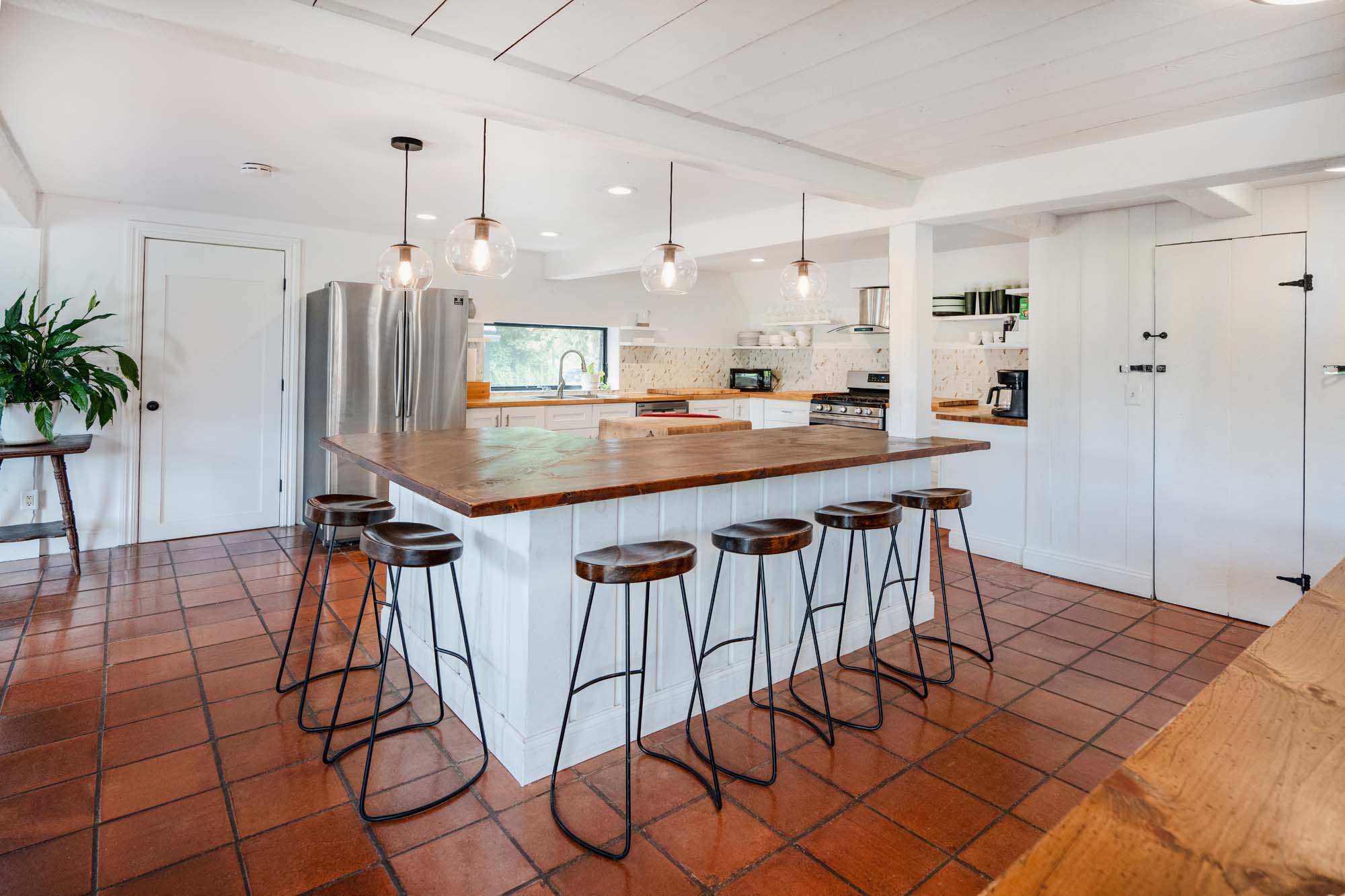 Modern kitchen with a large wooden island in the center surrounded by six high stools with wooden seats and black metal legs. The floor is covered with reddish-brown tiles. There are four pendant lights with glass globes hanging above the island. The kitchen has white cabinetry, a stainless steel refrigerator, a window above the sink, a stove, and various kitchen appliances and utensils. A green plant is on a small table to the left.
