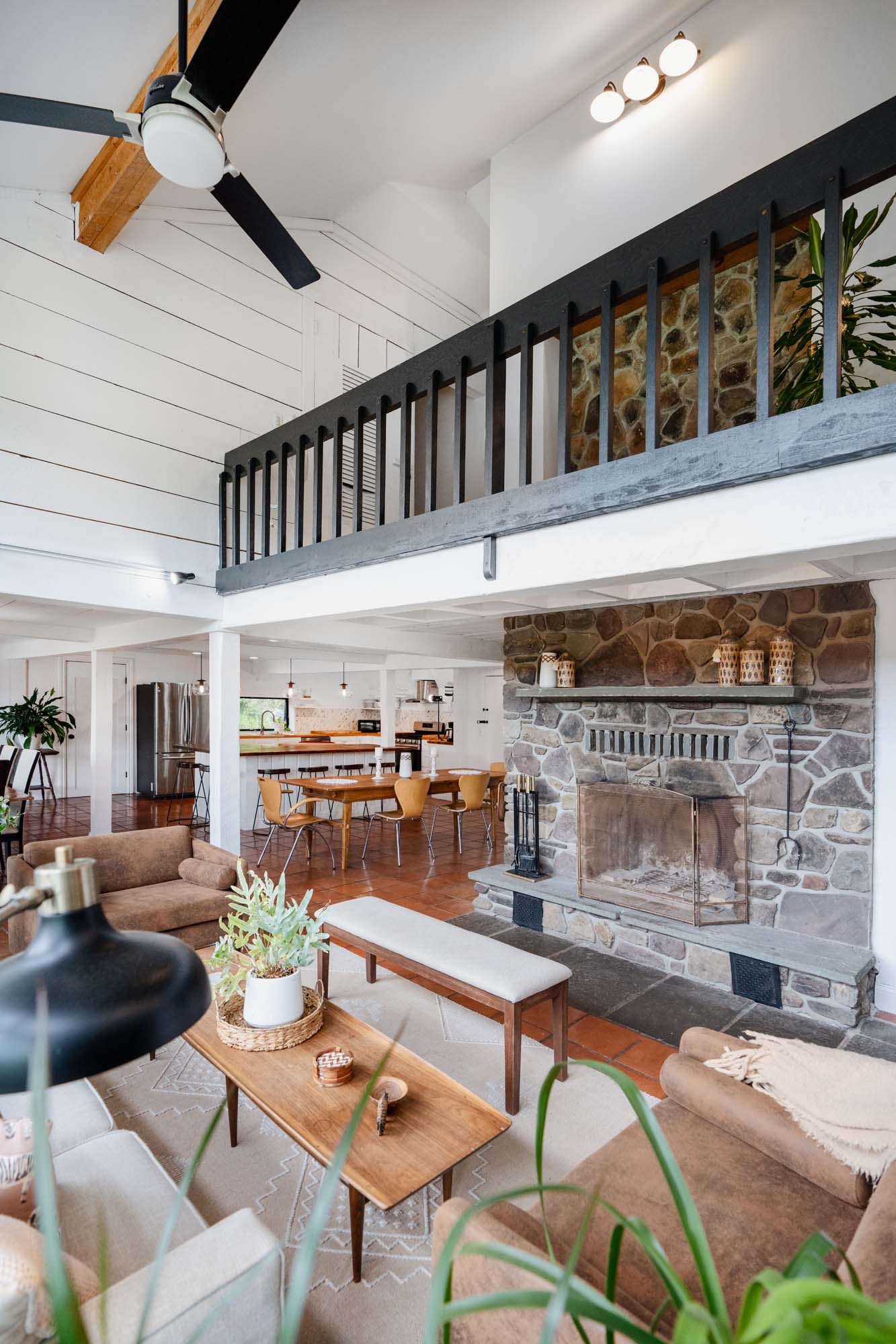 Interior living and dining area with a stone fireplace on the right side. The room features a high ceiling with a black ceiling fan and wooden beams. There is a wooden coffee table with a plant and decor items, surrounded by beige and brown seating. In the background, a dining table with six chairs is visible, along with a kitchen area with a refrigerator and bar stools. A balcony with black railing overlooks the space.