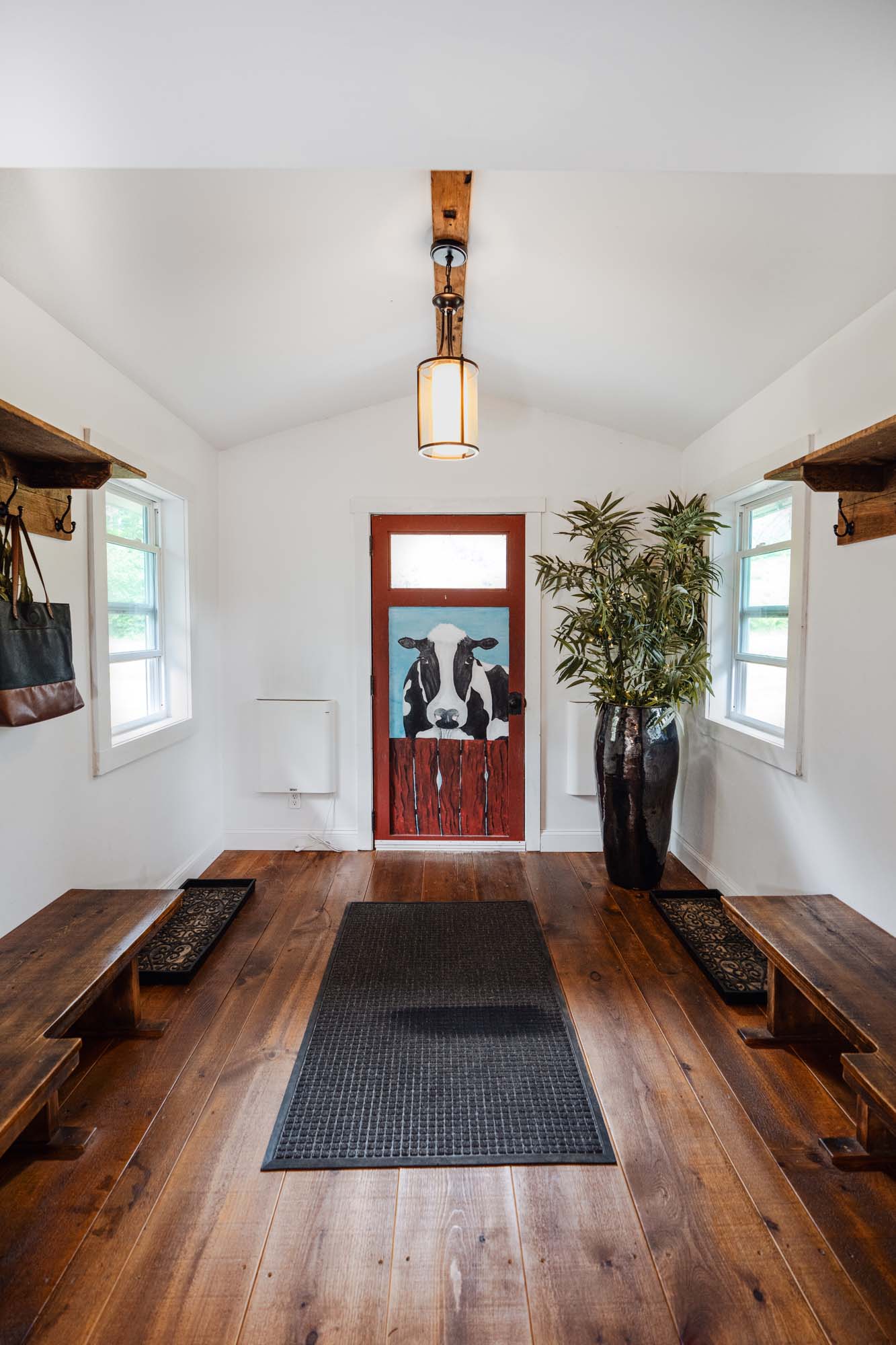 Small, rectangular room with white walls and a wooden floor. There is a door at the far end with a painting of a black and white cow on a blue background, partially obscured by a red fence. Two windows are on either side of the door. Wooden benches are placed along both walls, with black mats underneath. A large black vase with green leafy plants is on the right side near the door. A ceiling light fixture with a wooden beam is centered on the ceiling.